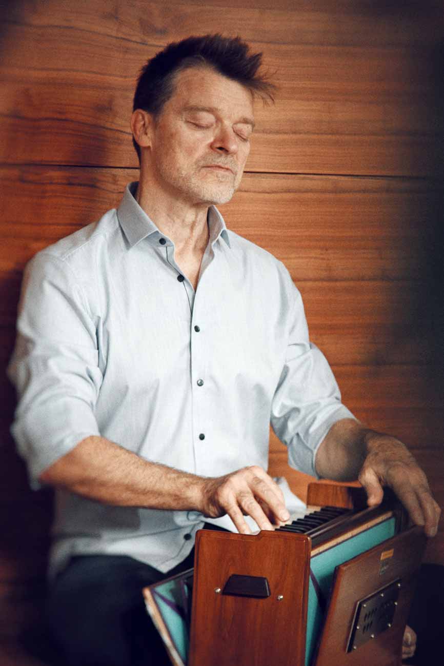 Jörg Buneru sitzt vor einer braunen Holzwand und spielt Harmonium Er hat die Augen geschlossen und trägt ein hellgraues, bis zum Ellebogen aufgekrempeltes Hemd.
