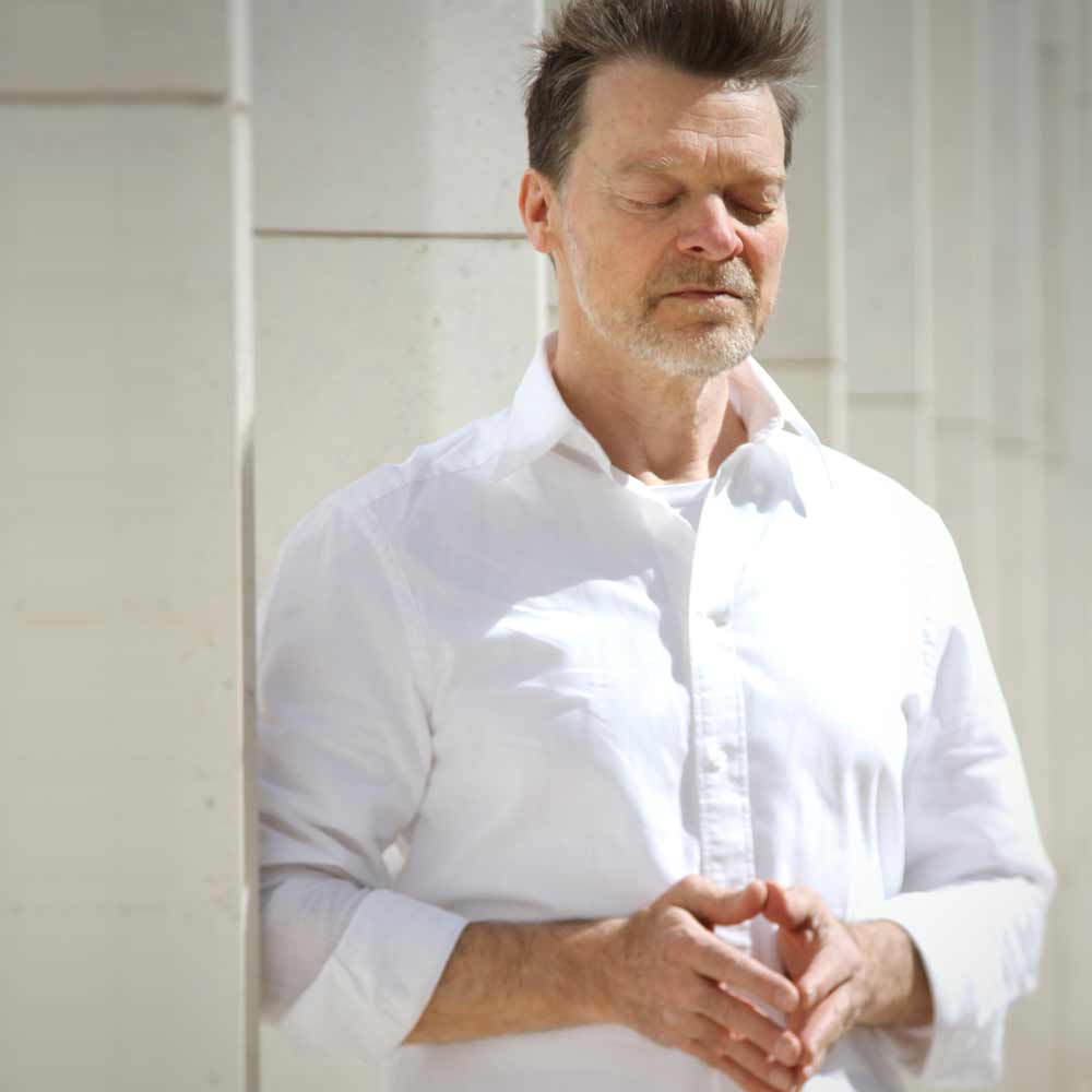 Porträtbild von Jörg Buneru, der vor einer hellgrauen Häuserfassade steht. Er hat die Augen geschlossen, die Fingerspitzen in Bauchhöhe aufeinander gelegt und die Augen geschlossen. Das Bild symbolisiert eine Achtsamkeitsübung und Meditation.
