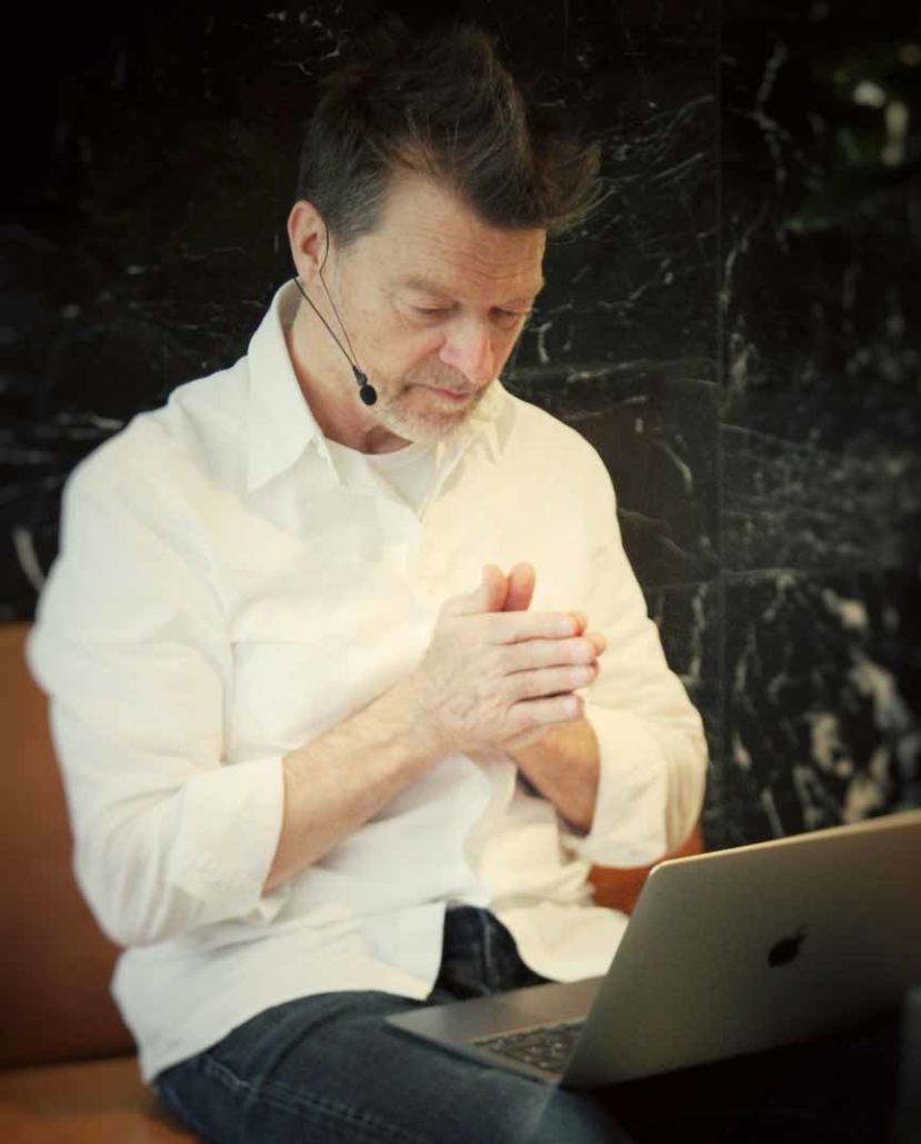 Jörg Buneru sitzt vor einem Laptop, der rechts im Bild erkennbar ist, und hat die Hände aneinander gelegt. Er trägt ein weißes, langärmeliges Hemd und ein Headset. Im Hintergrund eine schwarze Marmorwand. Das symbolisiert Jörg Bunerus Angebot zu Emotional Quality Coaching.